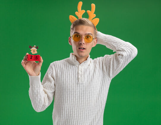 保持令人印象深刻的年轻帅哥戴着驯鹿鹿角头带戴着眼镜拿着圣诞树玩具和日期保持手后面的头隔离在绿色的墙上印象深刻圣诞节驯鹿