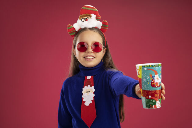 纸小女孩穿着蓝色高领毛衣 打着红色领带 头上戴着有趣的圣诞戒指 露出五颜六色的纸杯 快乐而积极的微笑小蓝色圣诞节