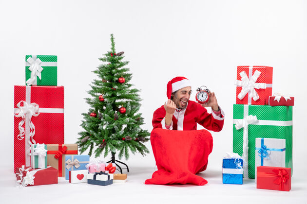 礼物表情滑稽的年轻人坐在地上庆祝圣诞节 在礼物和圣诞树旁展示钟表展示男人年轻人