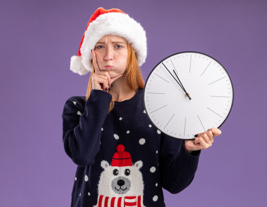 钟悲伤的年轻漂亮的女孩穿着圣诞毛衣和帽子拿着挂钟把手放在脸颊上隔离在紫色的背景上圣诞节脸颊毛衣