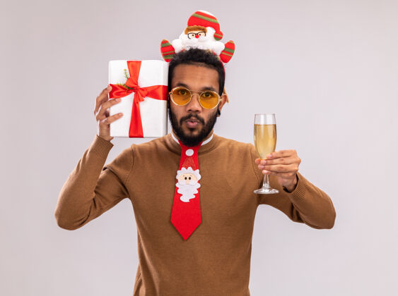 香槟一个非洲裔美国人 穿着棕色毛衣 头戴圣诞老人戒指 打着有趣的红色领带 手里拿着一份礼物 手里拿着一杯香槟 站在白色背景下惊讶地看着摄像机抱着非洲美国人