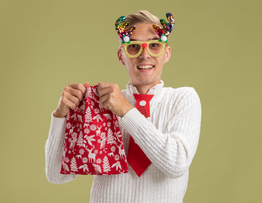 英俊快乐的年轻帅哥戴着圣诞新奇眼镜拿着圣诞袋准备打开它隔离在橄榄绿的墙上开放准备穿着