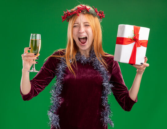 穿着兴奋的年轻漂亮的女孩穿着红色连衣裙 脖子上戴着花环和花环 手里拿着一杯香槟 绿色的墙上隔着一个礼盒玻璃杯圣诞节花环