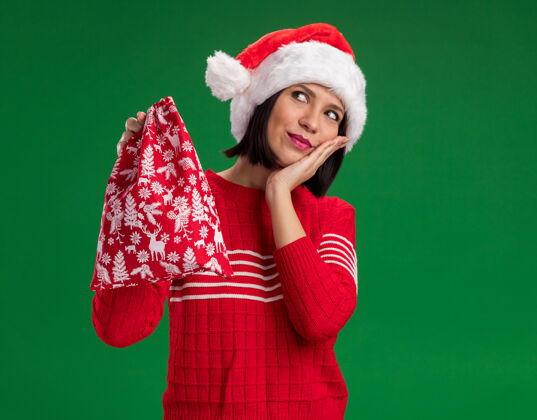 圣诞老人高兴的年轻女孩戴着圣诞帽拿着圣诞礼物袋手放在脸上看着绿色背景与复制空间隔离手礼物高兴
