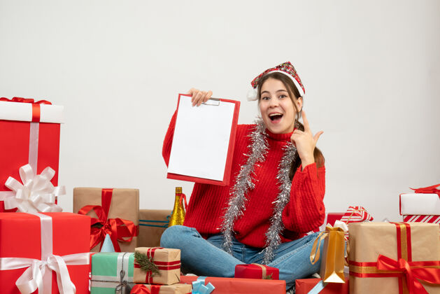 礼物戴着圣诞帽的快乐女孩拿着文件坐在白色的礼物旁边圣诞老人周围圣诞节
