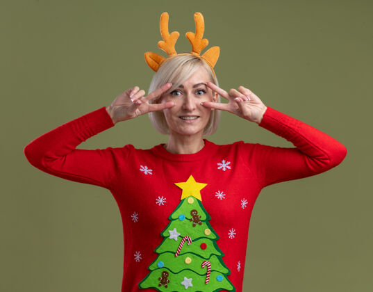 眼睛微笑的中年金发女人戴着圣诞驯鹿鹿角头带和圣诞毛衣看着相机显示的v符号在橄榄绿背景上孤立的眼睛附近看圣诞节相机