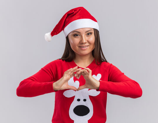 毛衣笑容满面的年轻亚洲女孩戴着圣诞帽和毛衣 在白色背景上显示出孤立的心形圣诞节女孩微笑
