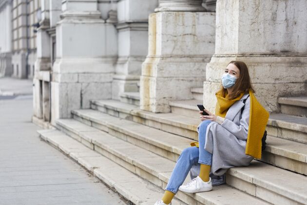 智能手机戴着医用面罩坐在台阶上用智能手机的女人水平Ncov病毒