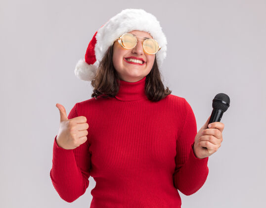 微笑穿着红色毛衣 戴着圣诞帽 戴着眼镜 拿着麦克风 兴高采烈地微笑着看着镜头 站在白色背景上竖起大拇指女孩站着拇指