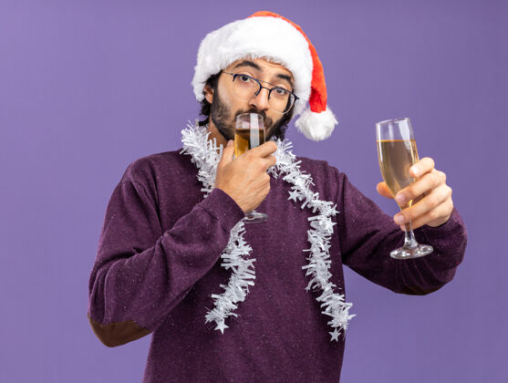 香槟高兴的年轻帅哥戴着圣诞帽 脖子上戴着花环 手里拿着一杯香槟和饮料 背景是蓝色的花环年轻抱着