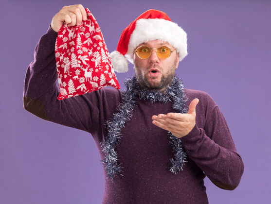 帽子令人印象深刻的中年男子戴着圣诞帽 脖子上戴着金箔花环 戴着眼镜 拿着并指着圣诞礼物袋 看着紫色背景上孤立的摄像机中年印象深刻礼物