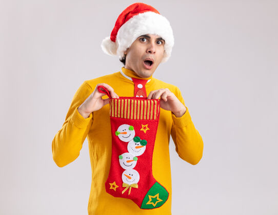 站着一个穿着黄色高领毛衣 戴着圣诞帽 打着滑稽领带的年轻人 手里拿着圣诞长袜 站在白色背景上 惊讶地看着镜头抱着搞笑惊讶