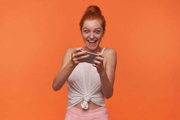 摆姿势室内拍摄喜出望外的年轻女性 披着白色上衣和粉色裙子的狐狸精发髻 手拿手机在橙色背景上摆姿势 脸上带着兴奋的表情在智能手机上玩游戏微笑抱着头发