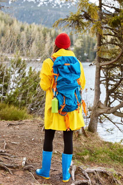 徒步旅行女游客背靠镜头 身着休闲黄色雨衣 胶靴 在山湖附近呼吸新鲜空气 引领活跃的生活方式背包森林山