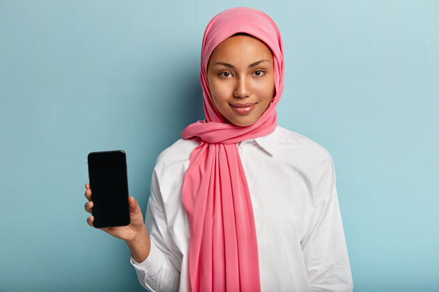 应用程序亚洲黑皮肤女性 戴粉色围巾 身穿白色衬衫 手持带有模拟屏幕的手机插入图像或文本 隔离在蓝色墙壁上选择性聚焦技术 文化 广告应用程序女人微笑