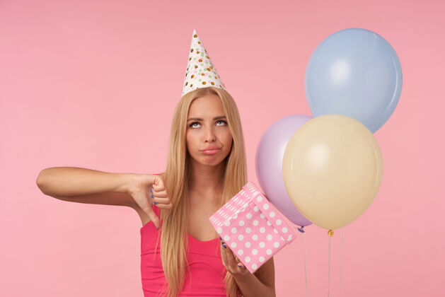 情感幻想破灭的长发金发女人 随意地展示她的拇指和滚动的眼睛失望 庆祝生日五彩气球 孤立在粉红色的背景长发圆锥体头发