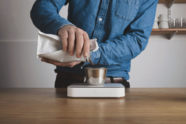 杯子一步一步压榨咖啡身着蓝色牛仔裤衬衫的咖啡师把烤豆从袋子里倒到白色砝码上的钢杯里专业咖啡冲泡咖啡店准备温暖的浓缩咖啡