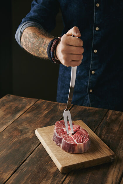 肉纹身的屠夫手拿着肉叉在新鲜的生牛排上当晚餐 面目全非烟美味屠宰