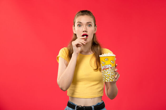 电影正面图年轻女性在电影院拿着爆米花包在浅红墙电影院看女性趣味电影灯光艺人爆米花
