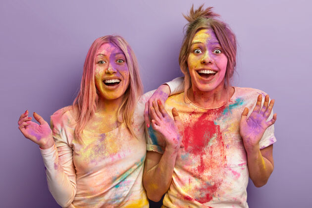 肖像胡里节 有趣和人的概念两个欣喜若狂的女人玩颜色 展示手掌 涂上五颜六色的粉末 孤立在紫色的墙壁五颜六色爆炸胡里积极庆祝