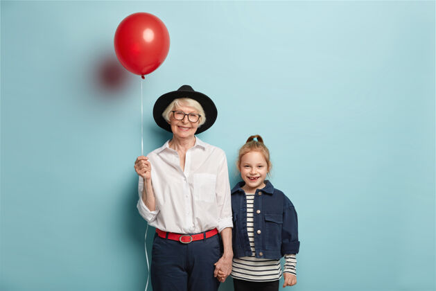 头饰微笑的红发女孩带着母亲节来祝贺奶奶 穿着条纹套头衫和牛仔夹克高兴的老太太戴着时髦的黑色帽子 拎着气球 牵着小孙女的手时尚奶奶奶奶