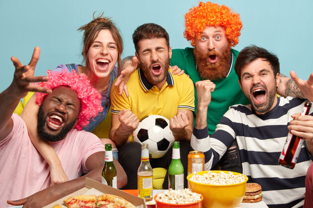啤酒男女球迷在家里看电视上的足球 享受刺激的比赛 握紧拳头 庆祝胜利 表达积极的情绪 在碗里吃爆米花 吃比萨饼 在蓝色的墙上摆姿势比赛多样化女人