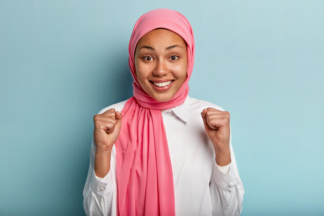 衣服祈祷民族妇女举起拳头 欢庆胜利 满意工作的好结果 戴着粉色头巾 穿着白衬衫 隔着蓝色的墙手势幸福姿势民族东方