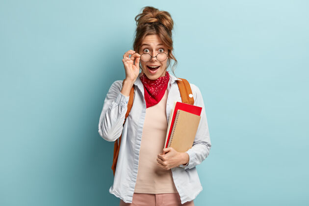风格惊喜快乐的女生透过眼镜看 手放在相框上 拿着螺旋笔记本和红本子大学教育学习