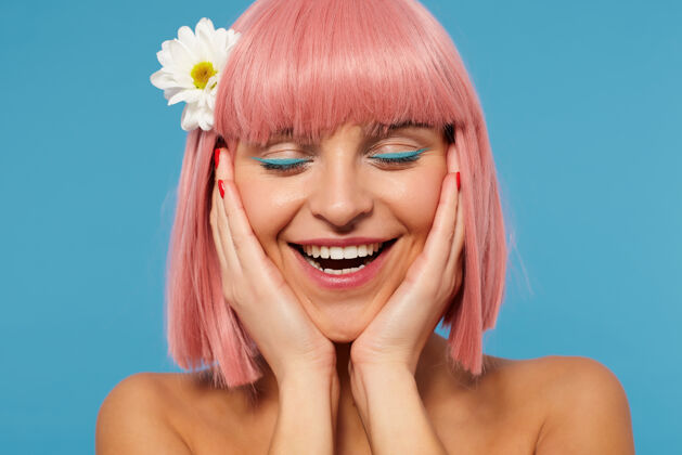 脸特写镜头：可爱的年轻积极的粉红色头发的女士举着她的脸与提高手掌 同时微笑着愉快地闭上眼睛 站在蓝色的背景下肖像情感眼影