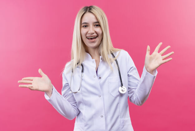 粉红色微笑的医生年轻的金发女孩穿着医用长袍戴着听诊器和牙套在孤立的粉红色背景上摊开双手穿着支撑金发