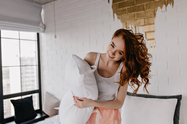 年轻搞笑的高加索女士拿着枕头在早晨嬉戏地摆姿势优雅的欧洲卷发女孩在享受拍照漂亮红发快乐