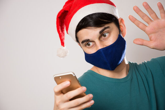 圣诞老人年轻的西班牙裔男子戴着面具 戴着圣诞老人帽 一边打视频电话冠状病毒手机冠状病毒19