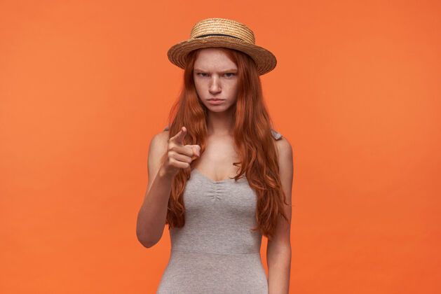 女士愁眉不展的年轻红发女子的肖像 留着一头长发 身穿灰色衬衫 头戴草帽 在警告牌上竖起食指 在橙色背景上摆姿势脸波浪沉思