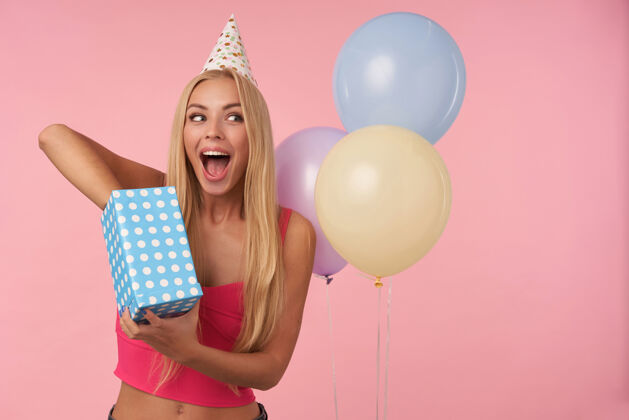 上衣快乐的年轻金发女性 长发在粉色背景下摆姿势 打开礼物 对收到生日礼物感到兴奋和惊讶人 娱乐和节日属性室内头发长发