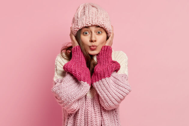 针织欧洲女人的画像戴着针织头饰 穿着宽松的毛衣 想亲吻某人 隔着粉色的墙壁休闲风格冬季时尚概念人类的面部表情时尚成人面部