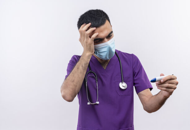 面罩吓得年轻的男医生穿着紫色手术服 戴着听诊器医用口罩 把手放在额头上 看着手边孤立的白色背景上的温度计紫色前额医生