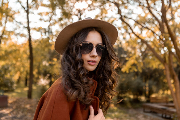 夹克迷人的时尚微笑的女人卷发漫步在街头公园穿着温暖的棕色外套秋季流行时尚 街头风格戴帽子和太阳镜装束优雅户外