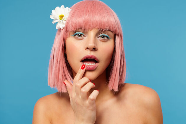 年轻室内拍摄的年轻漂亮的绿眼女性 粉色短发 用食指举着涂唇膏 赤裸着肩膀站在蓝色背景下20秒情感手
