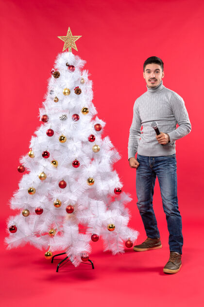 红色圣诞气氛 自信的家伙穿着牛仔裤站在装饰好的圣诞树旁 拿着麦克风 表情惊讶心情圣诞自信的男人
