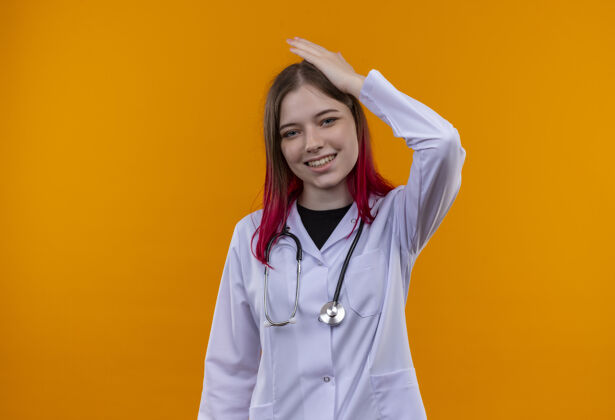 橙色微笑的年轻医生女孩穿着听诊器医用长袍 手放在头上 孤立的橙色背景医生微笑头