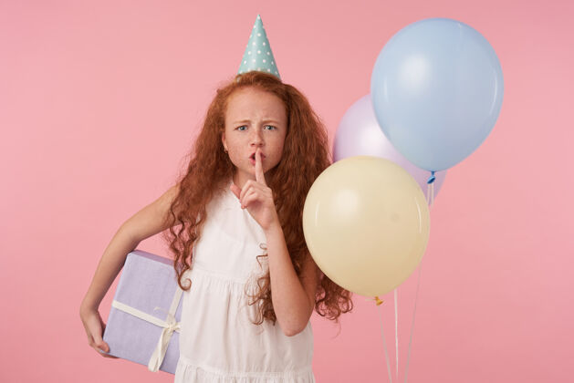 女孩严肃的小女孩 一头浓密的卷发 穿着节日的衣服 站在粉色的背景和彩色的气球上 手里拿着礼品盒 举起食指 要求保持沉默食指节日欧洲