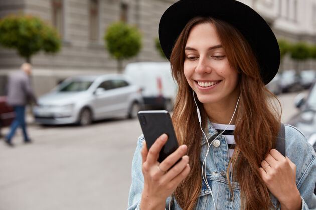 浏览戴着帽子的快乐女性 手拿手机 听播放列表中的音乐 在城市环境中闲逛短信社交在线
