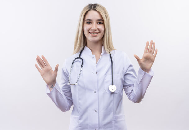 医疗微笑的医生年轻的金发女孩戴着听诊器和医用长袍戴着牙套在孤立的白色背景上举手提高牙科女孩