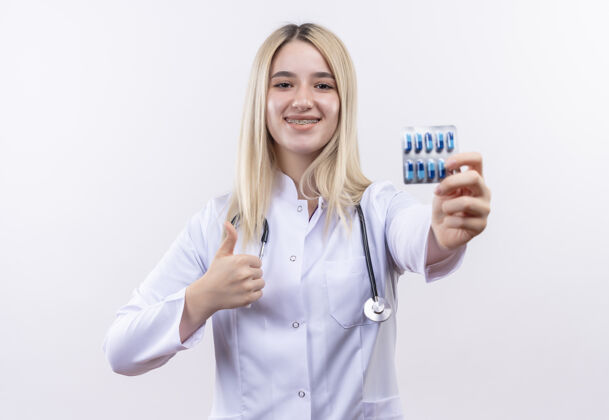 抱着微笑的医生年轻的金发女孩戴着听诊器和医用长袍 戴着牙套 拿着药片 在孤立的白色背景上竖起大拇指向上听诊器拇指