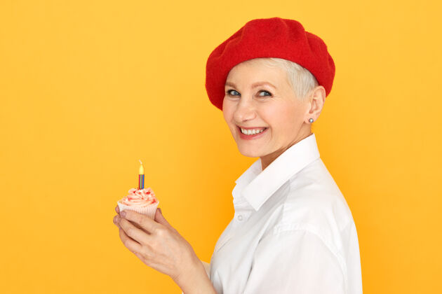 肖像红帽下染短发的中年女子手持生日纸杯蛋糕许愿的侧视图生日蜡烛时尚