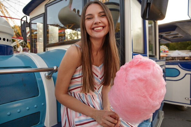 快乐美丽的年轻黑发女士的肖像 穿着浪漫的裙子 面带迷人的微笑 倚靠在假蒸汽火车上 手里拿着粉色的棉花糖肩带积极美丽