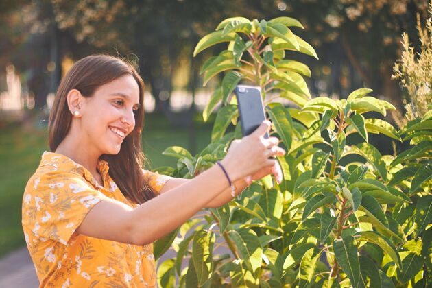 公园一个穿着黄色衬衫的年轻女孩在灌木丛旁自拍的美丽照片白天通讯手机