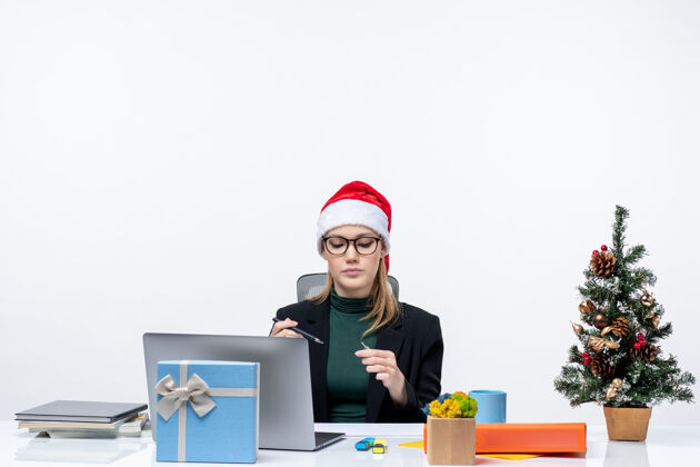 金发一个严肃的金发女人 戴着圣诞老人的帽子 坐在一张桌子旁 桌子上放着一棵圣诞树和一份白色背景的礼物笔记本电脑圣诞树办公室