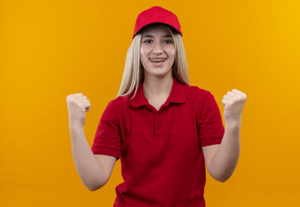 红色微笑的递送年轻女孩穿着红色t恤 戴着牙套 在孤立的橙色背景上显示“是”的手势女孩手势展示