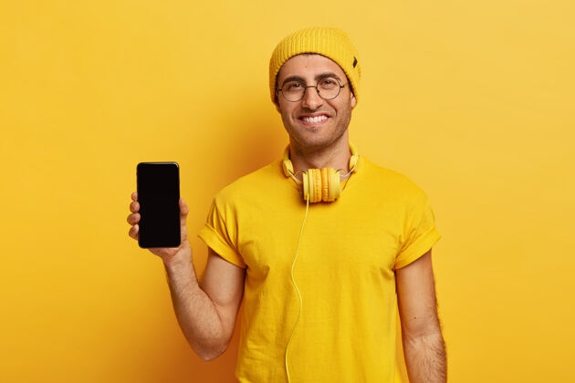 男性满意的欧洲男人拿着黑色的模拟空间的现代手机 戴着黄色的头饰音乐促销正面